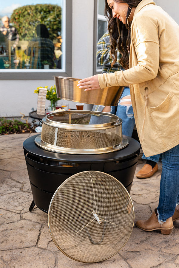 Person assembling a TIKI® Brand fire pit with a gold mesh screen and insert on a backyard patio.