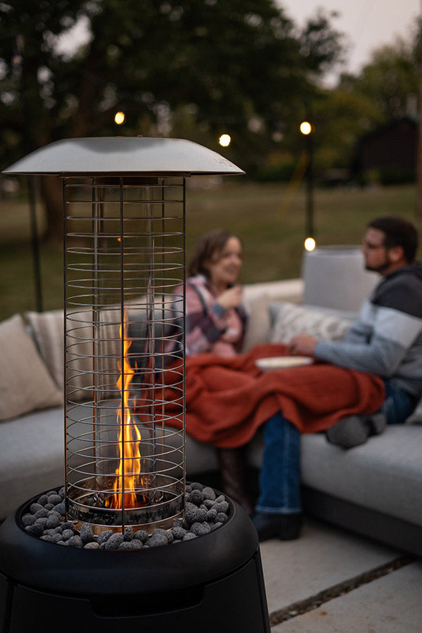 Outdoor patio heater with visible flame, warming two people relaxing on a sofa with blankets in a cozy backyard setting.