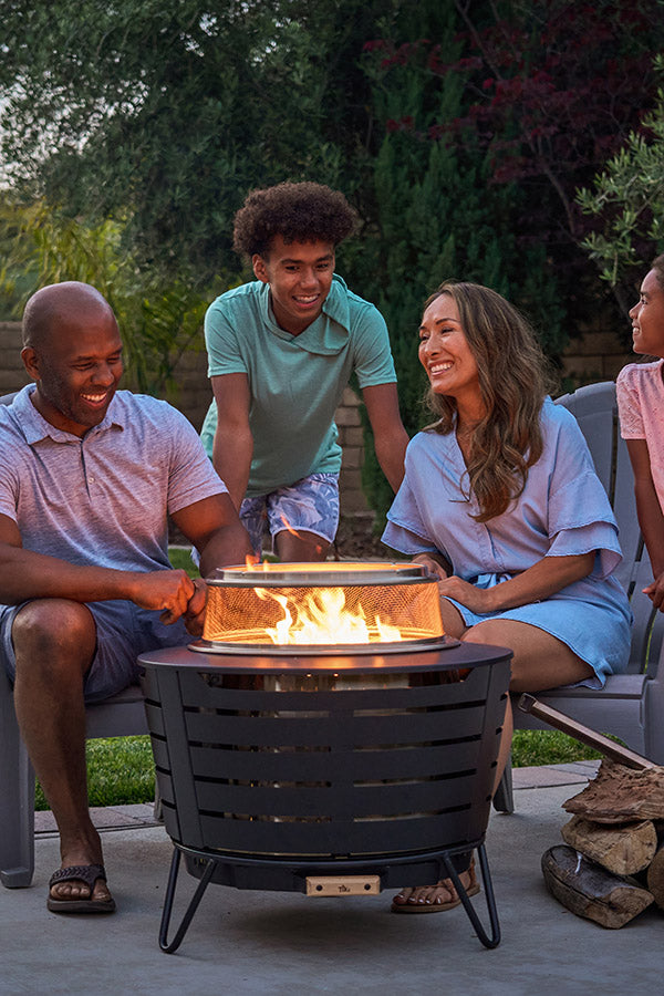 Family enjoys a cozy evening outdoors around a TIKI® fire pit, seated on patio chairs with firewood stacked nearby.
