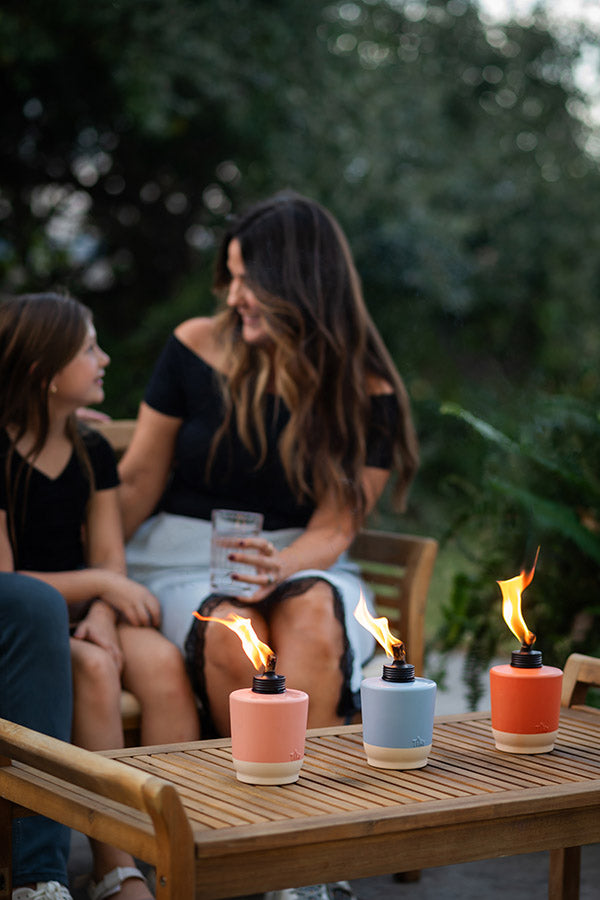 Three colorful TIKI tabletop torches with flames on a wooden patio table; people relaxing outdoors in the background.
