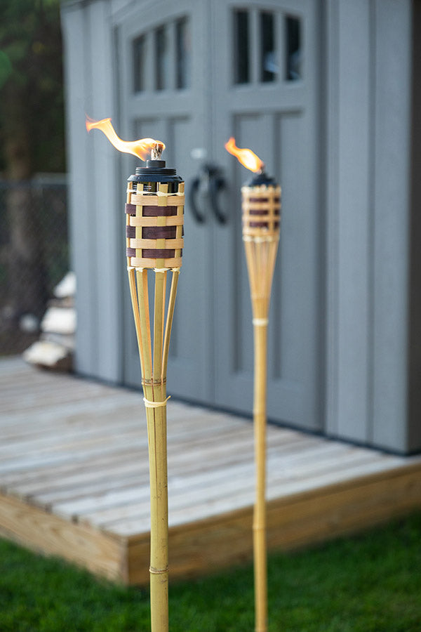 Two bamboo TIKI torches with flames lit, placed on grass near a wooden deck and gray shed.