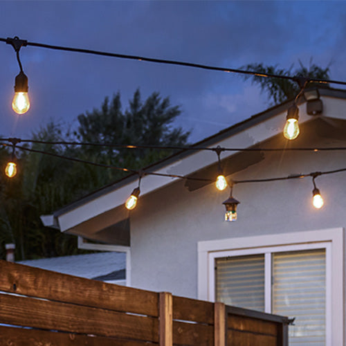 String lights glow above a backyard patio at dusk, creating a warm, inviting outdoor atmosphere near a house.