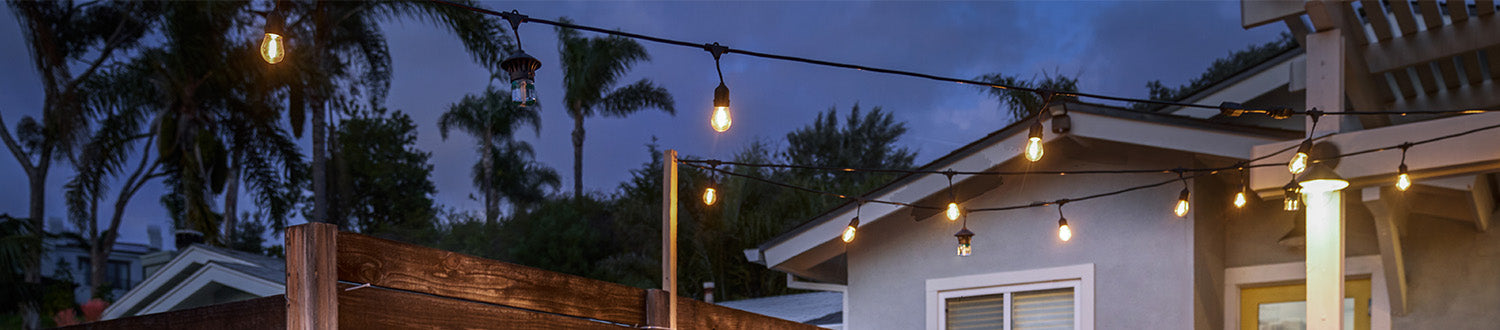 String lights illuminate a backyard patio at dusk, creating a warm, inviting outdoor space near a house and palm trees.