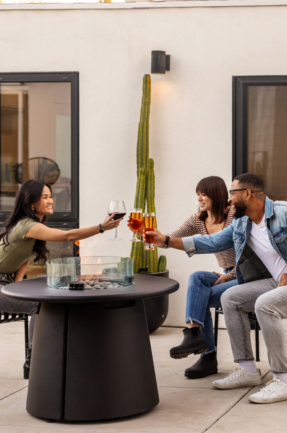 Three people toast drinks around a modern outdoor fire pit table on a patio, enjoying a cozy evening together.