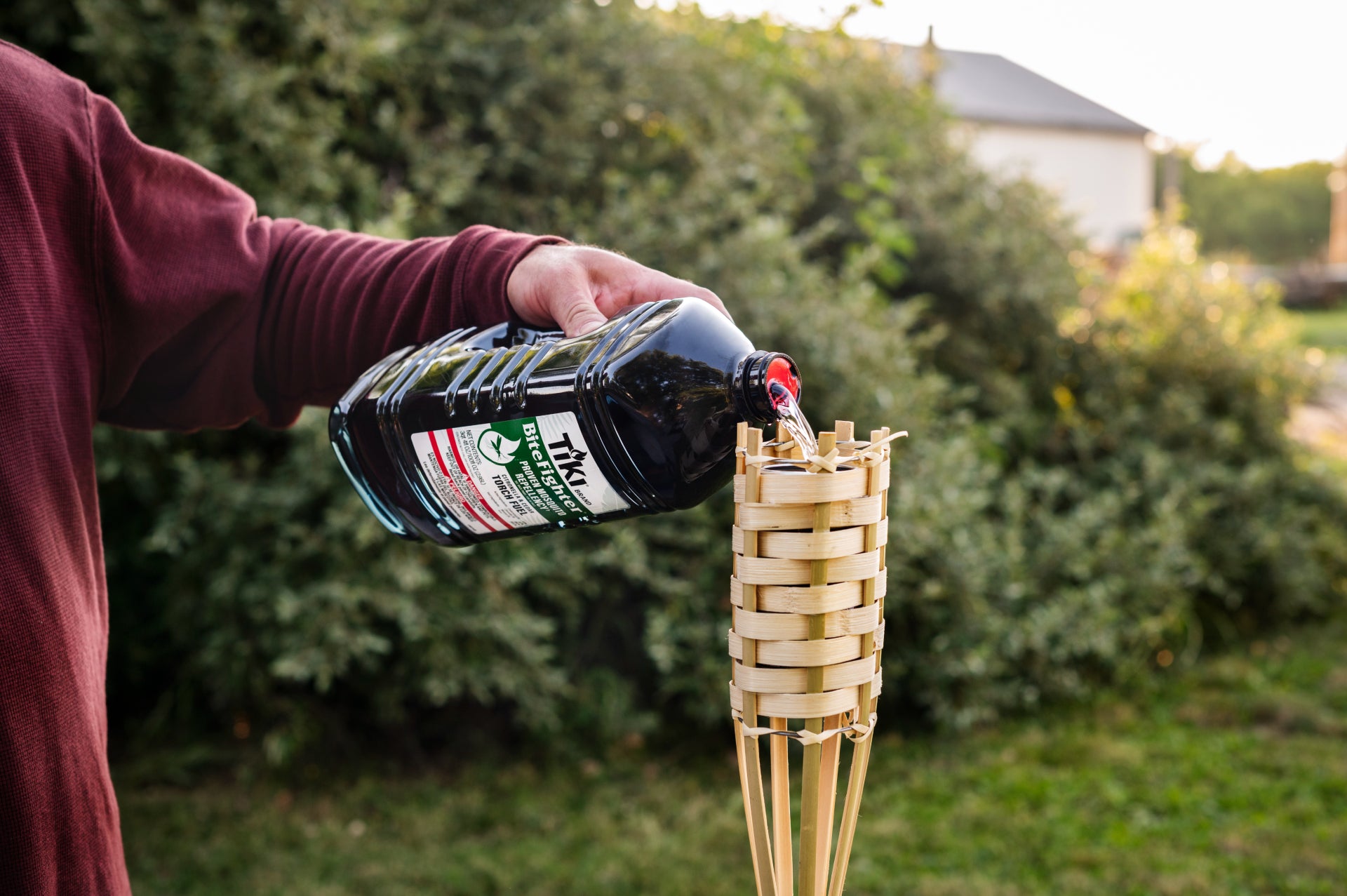 Person pours TIKI Brand BiteFighter torch fuel into a bamboo torch outdoors, preparing for mosquito protection.