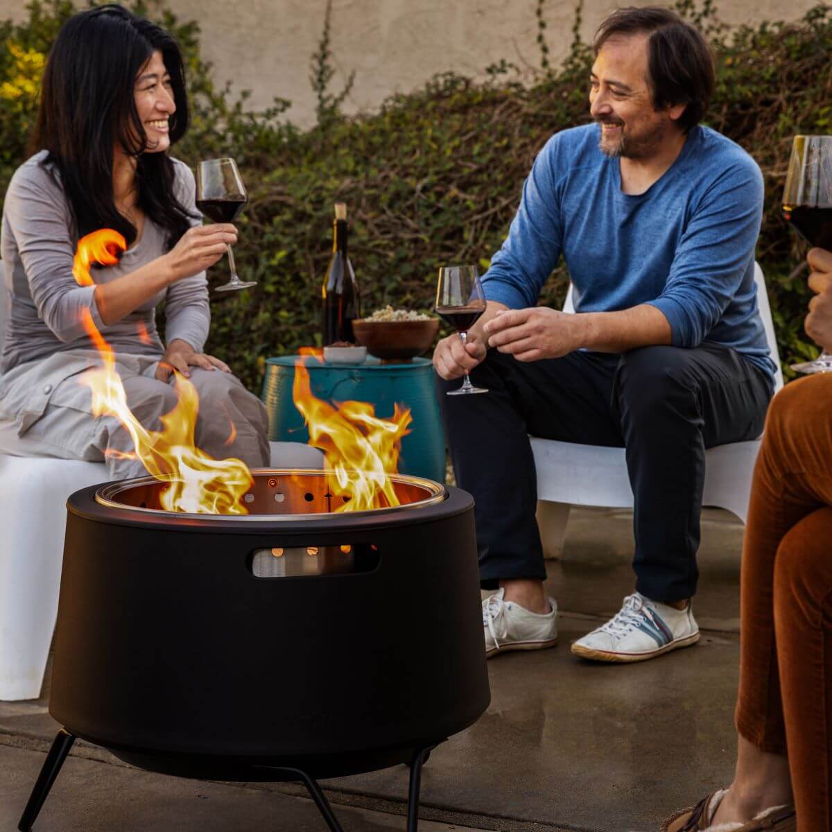 Black fire pit with flames, outdoor setting, people enjoying wine.