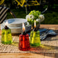 Set of three glass oil lamps in green and red with flames, on a table with plates and flowers.