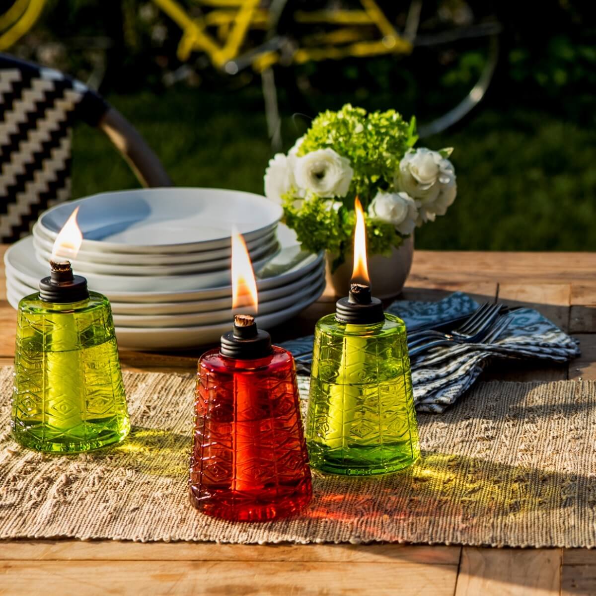 Set of three glass oil lamps in green and red with flames, on a table with plates and flowers.