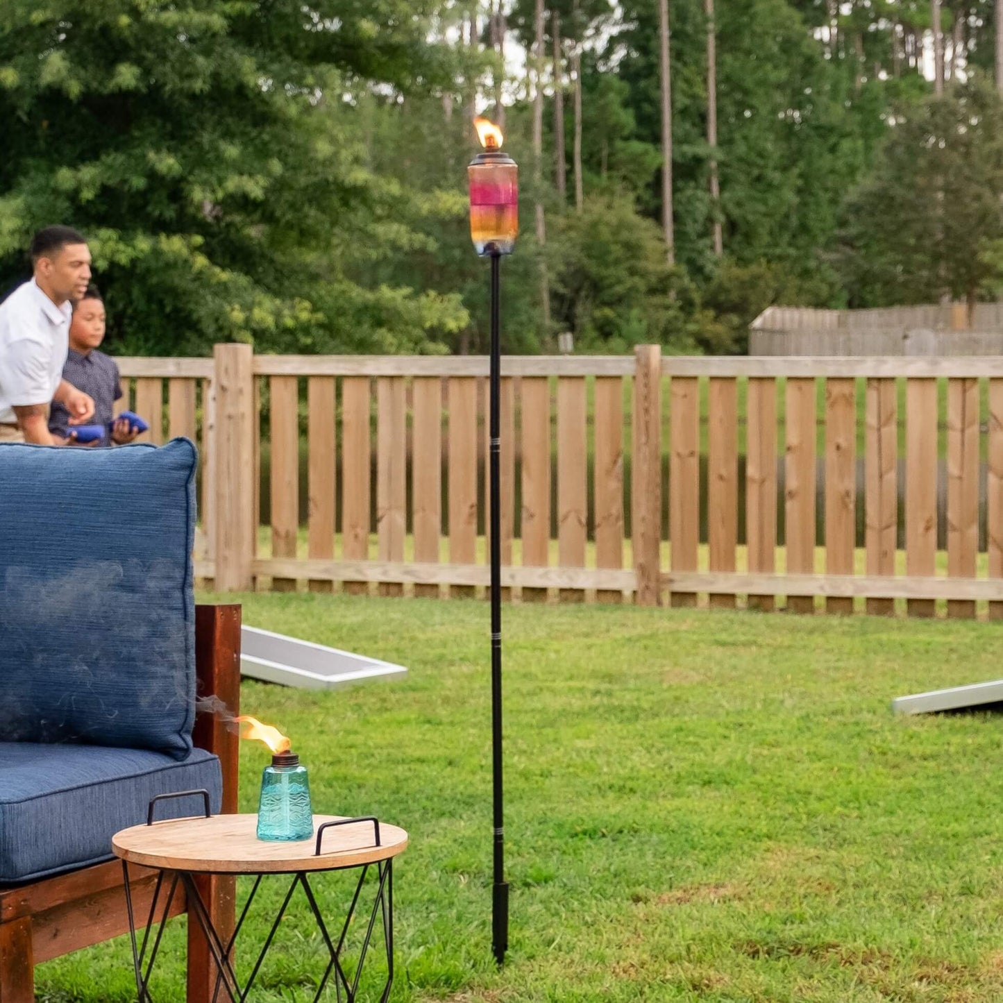 Glass tiki torch on a tall black pole, lit with a visible flame, set in a grassy backyard near outdoor seating.