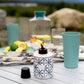 White Tiki torch with blue geometric pattern, lit flame, on outdoor table with cups and lemons.