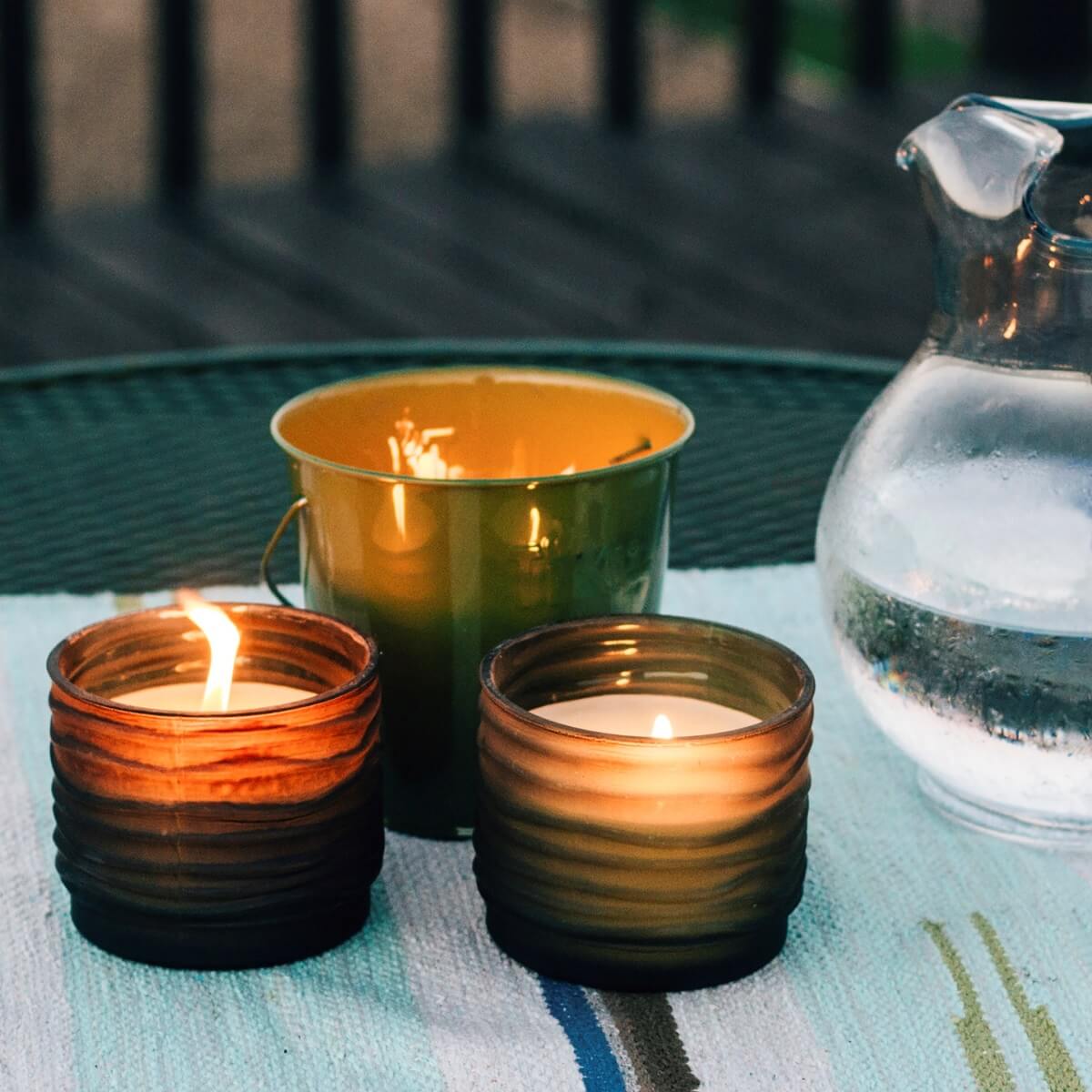 Three lit candles in amber glass containers on a table with a glass pitcher.