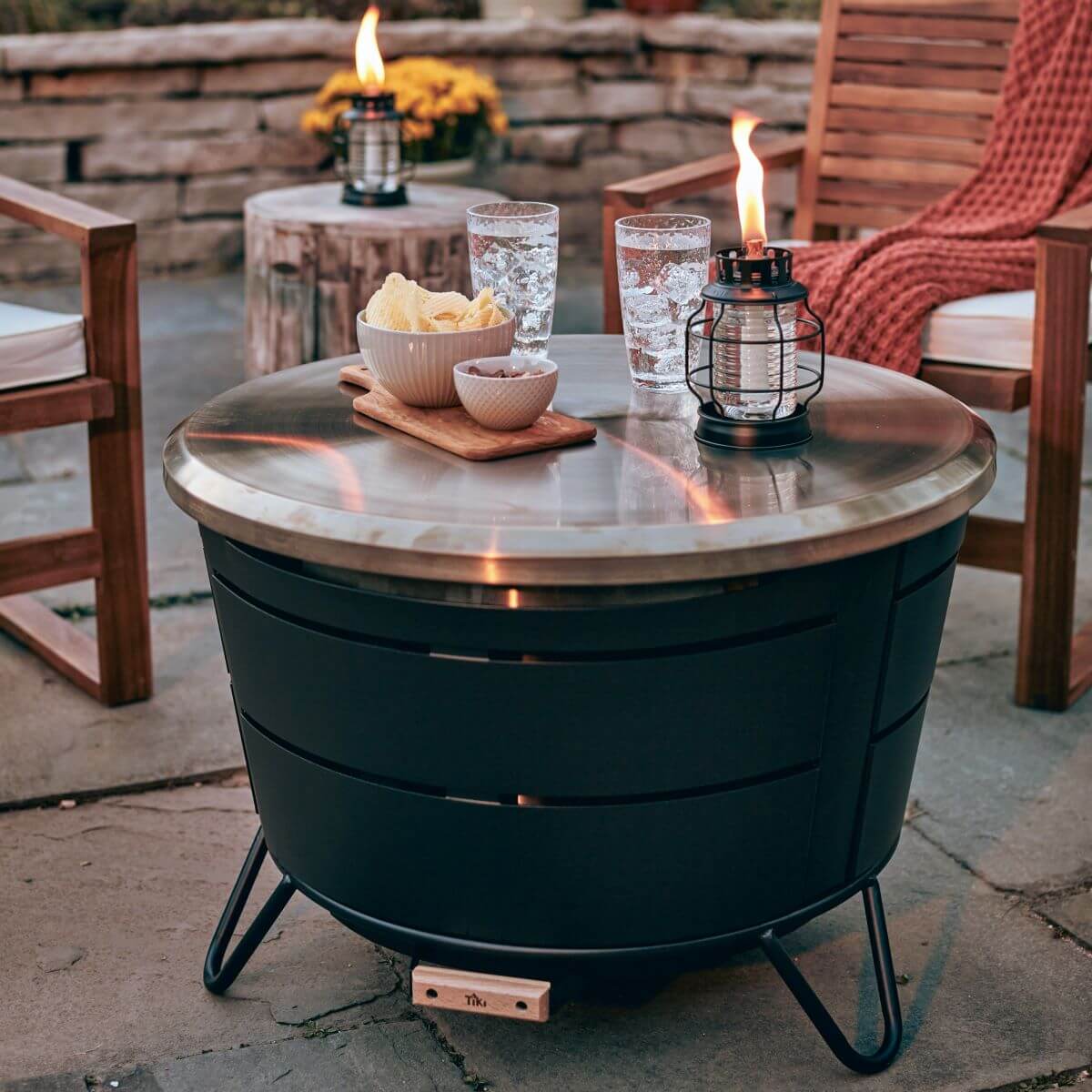 Black round fire pit table with glass top, lanterns, and snacks.
