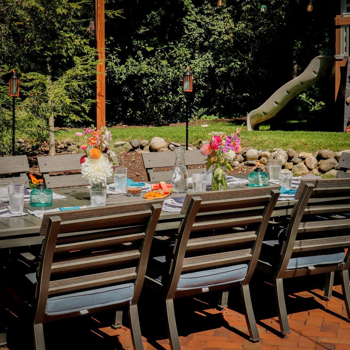 Outdoor dining set with wooden tables and chairs, colorful floral centerpieces, and glassware.