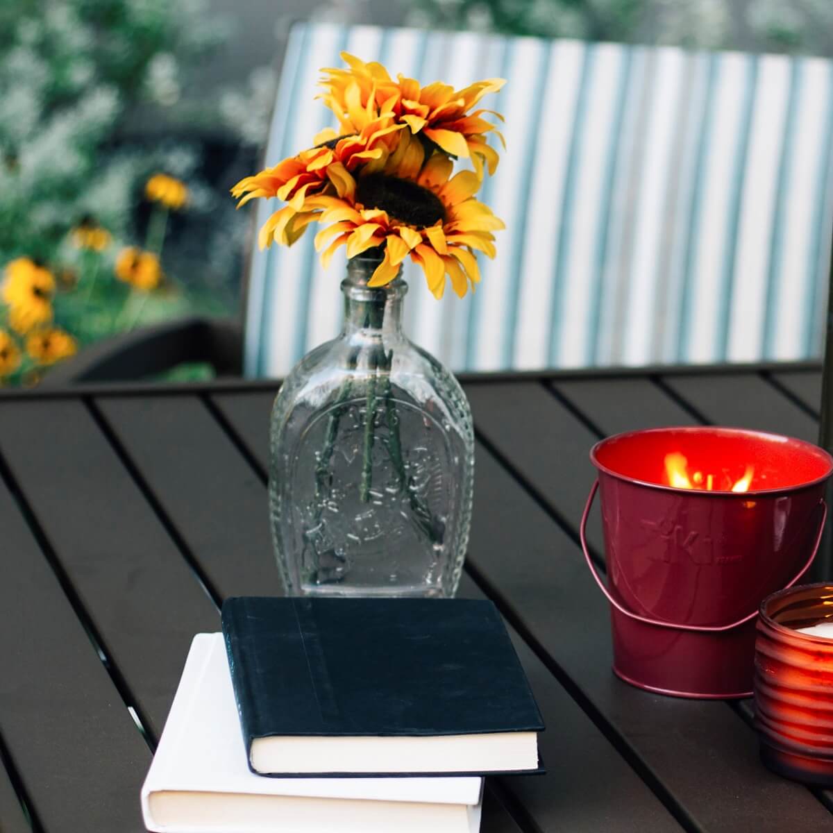 Clear glass vase with sunflower, red bucket candle, and stacked books on table.