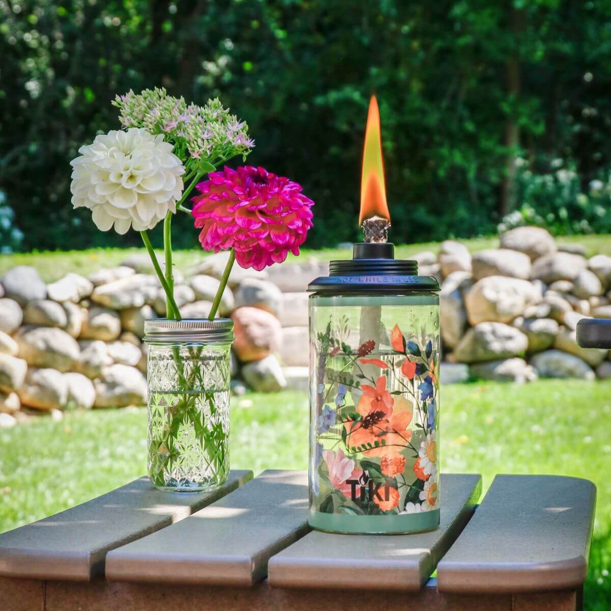 Tiki floral-patterned oil lantern with flame, beside a mason jar with white and pink flowers.