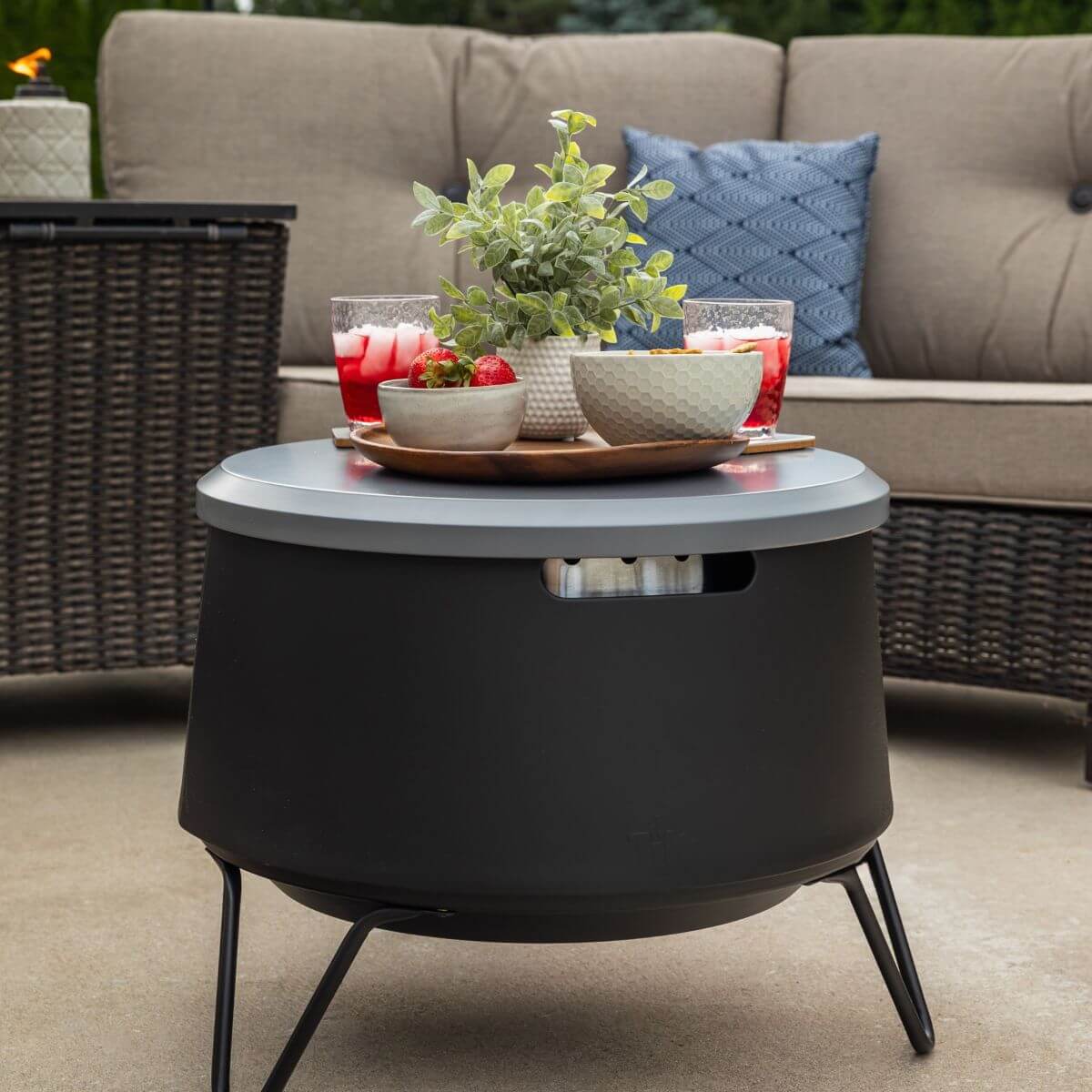 Black round outdoor fire pit table with gray top, surrounded by wicker furniture and decorative items.