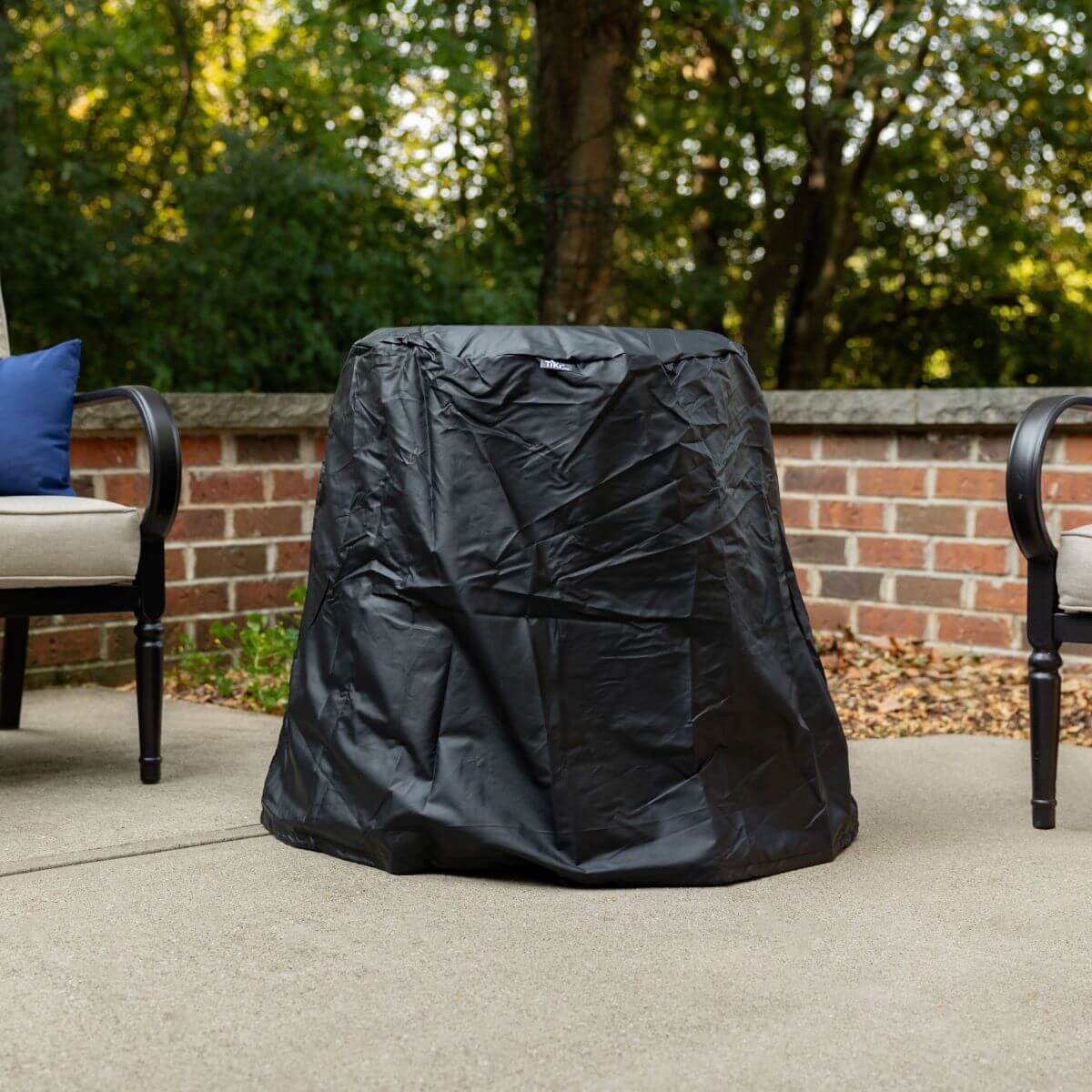 Black outdoor grill cover on patio with brick wall and trees in background.