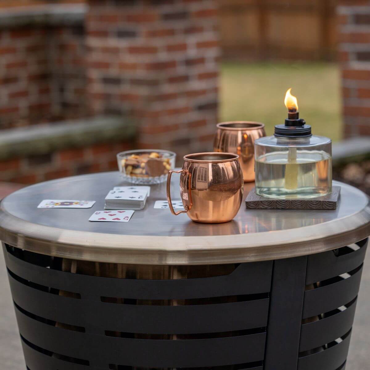 Outdoor patio table with copper mugs, playing cards, and a lit oil lamp.