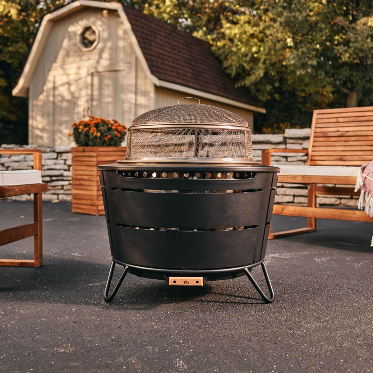Black TIKI outdoor fire pit with mesh cover, set on patio near wooden benches and shed.