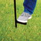 A person uses a black metal garden tool with a foot step to press into green grass for easy soil penetration.