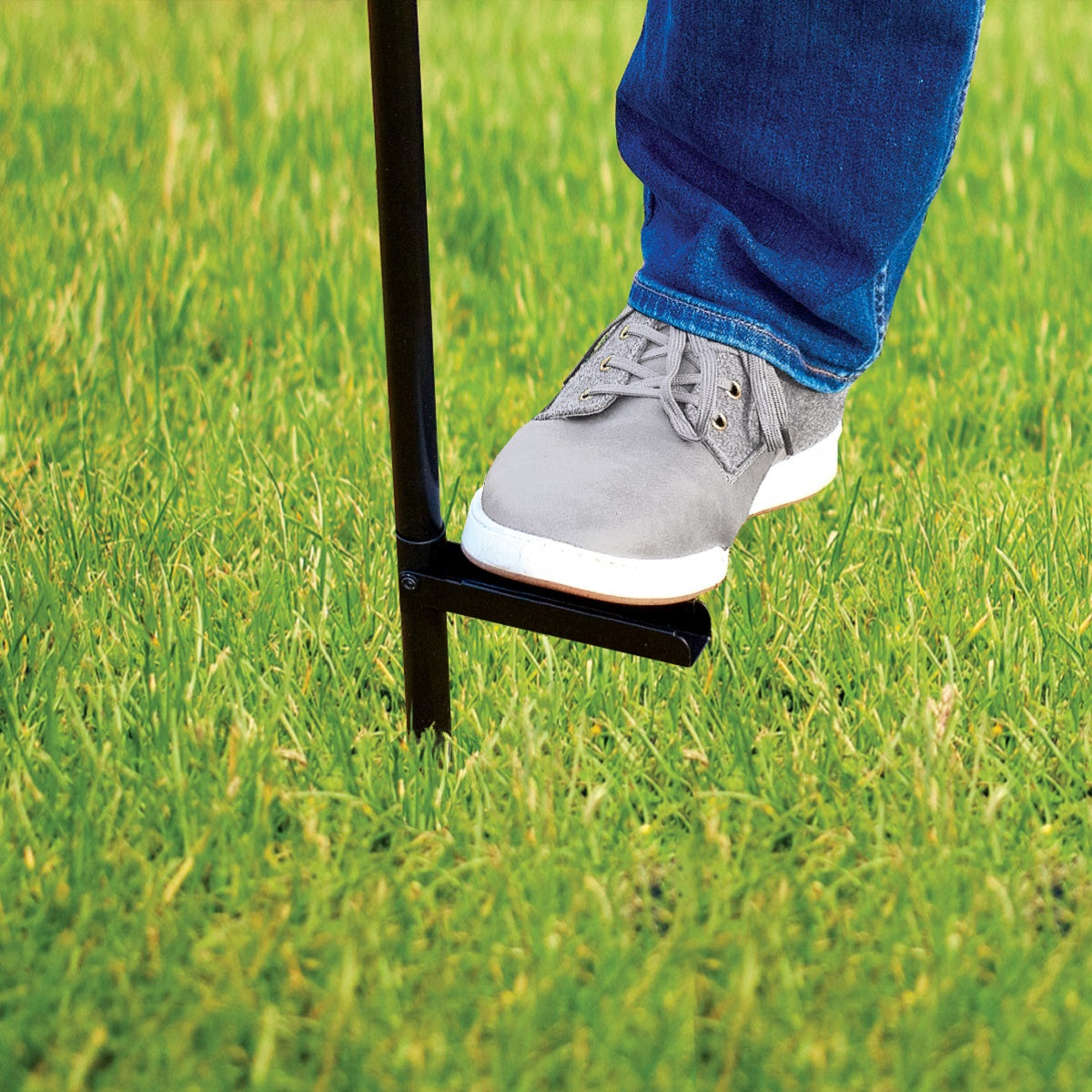 A person uses a black metal garden tool with a foot step to press into green grass for easy soil penetration.