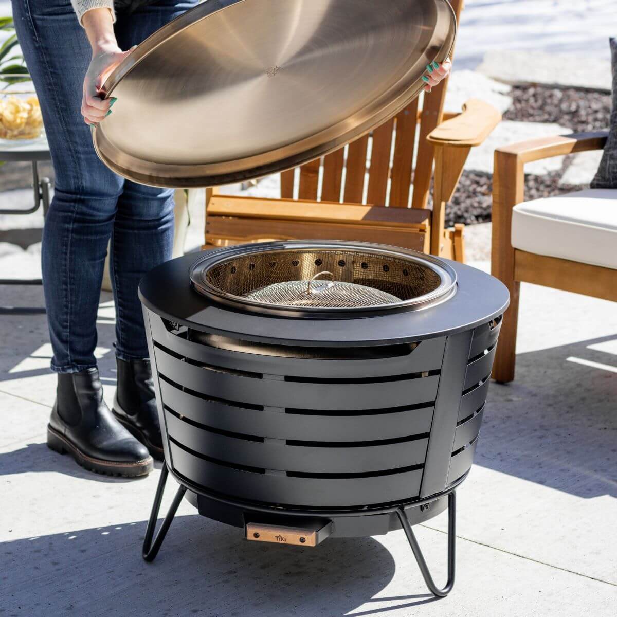 Gray outdoor fire pit with lid being lifted, showing grill grate inside.