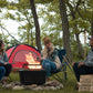 Black portable fire pit with glowing flames, surrounded by campers near a red tent.