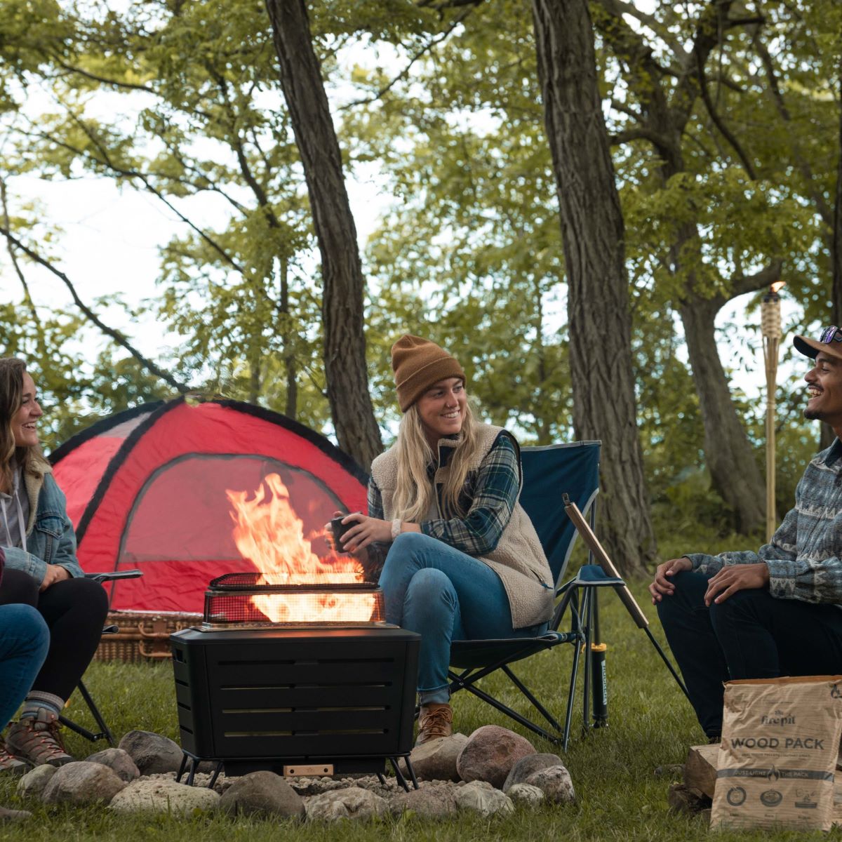 Black portable fire pit with glowing flames, surrounded by campers near a red tent.