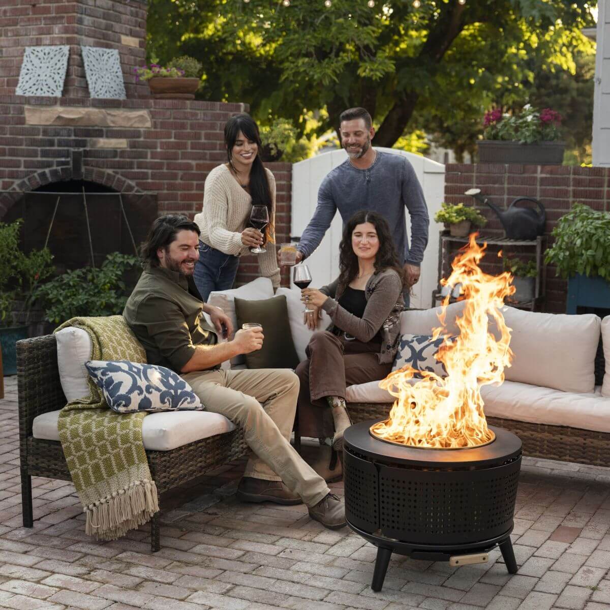 Black outdoor fire pit with roaring flames, surrounded by cozy patio furniture and people enjoying drinks.