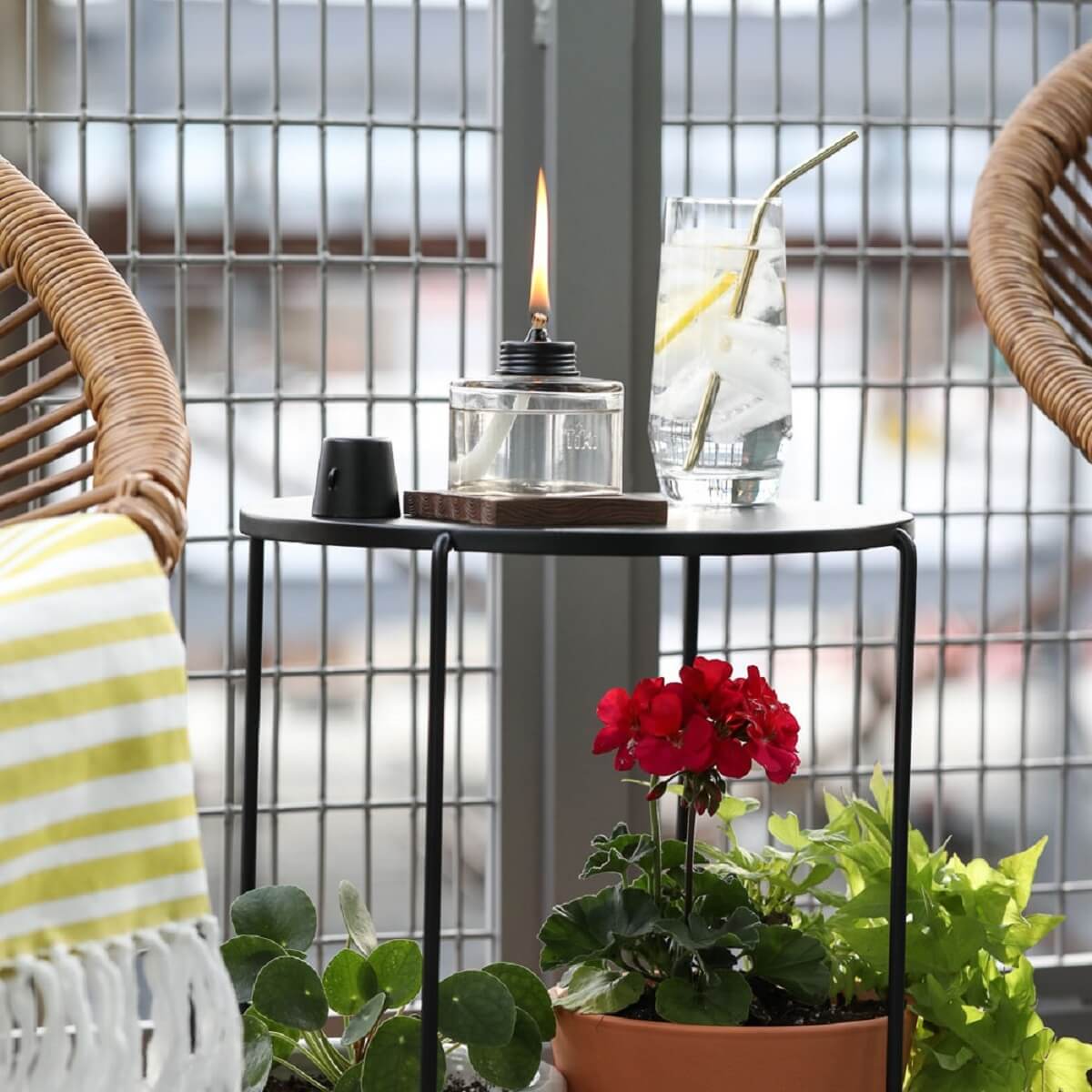 Clear glass oil lamp with black cap, lit flame, on black metal table with drink and plant.