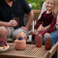 Terracotta tabletop lantern with flame, red cups, and snack bowl on wooden table.
