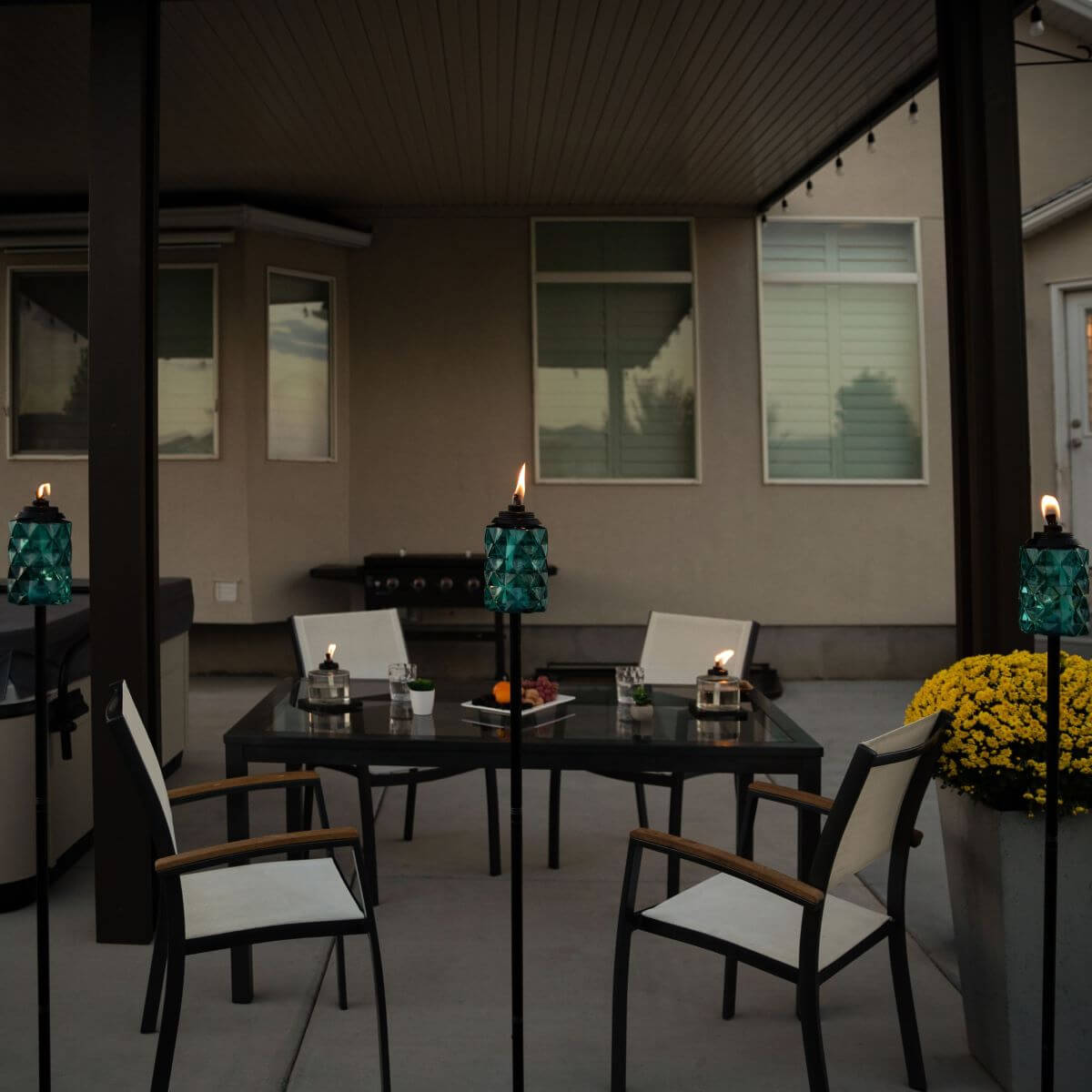 Outdoor patio set with glass table, white chairs, and teal tiki torches. Modern, accessible seating and decor.