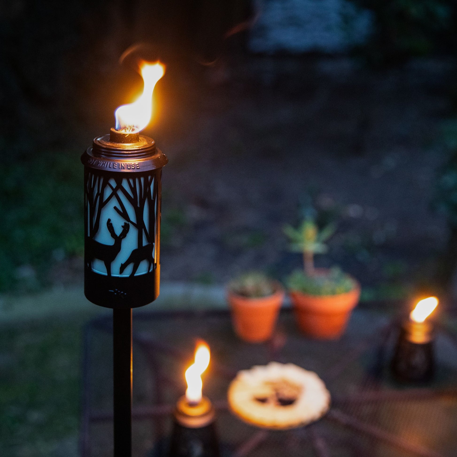 Black tiki torch with deer design, lit flame, outdoor setting.