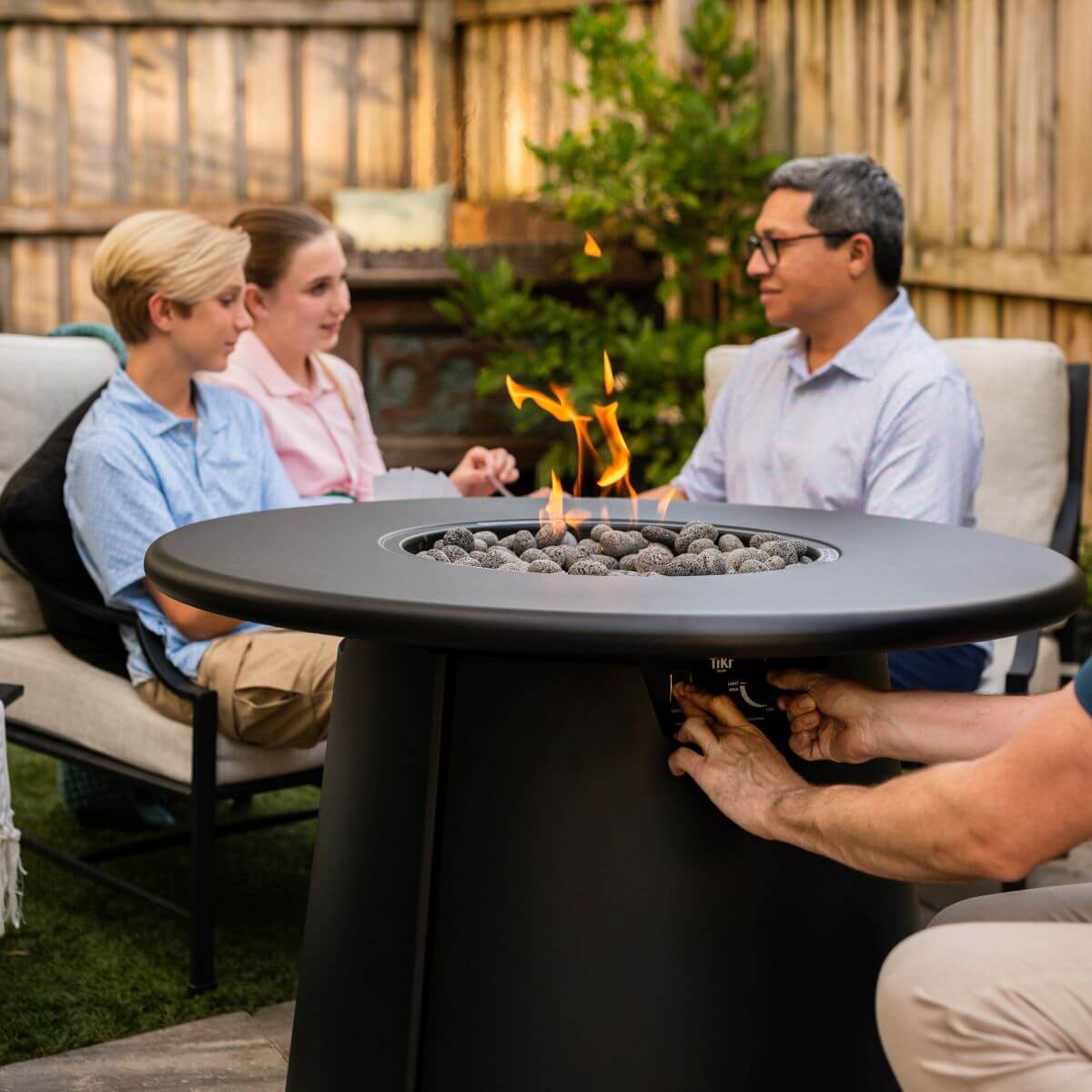 Black Tiki fire pit table with flames and stones, surrounded by people outdoors.