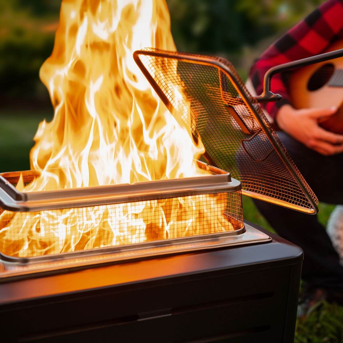Portable fire pit with mesh lid and flames, red plaid shirt in background.