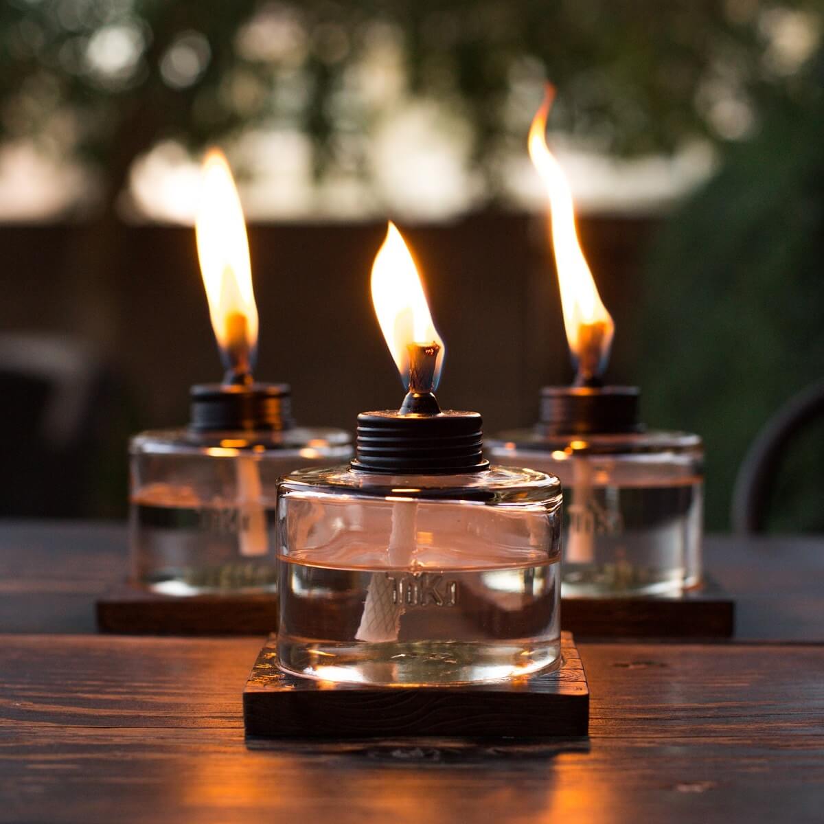 Clear glass oil lamps with black tops, lit flames, on wooden bases.