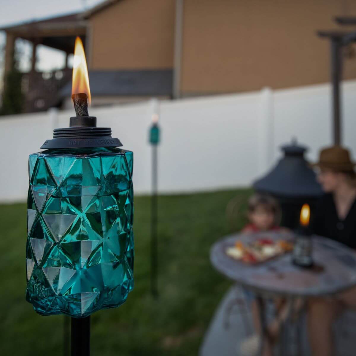 Blue glass outdoor tiki torch with geometric pattern, black top, and visible flame, set in a backyard garden.