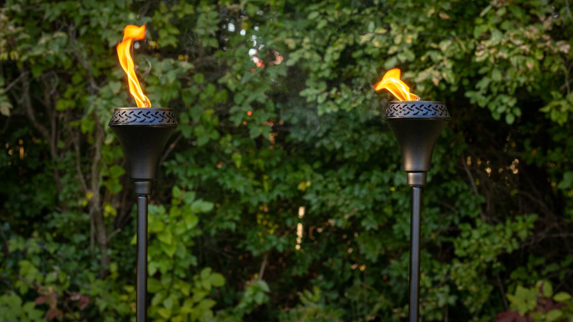 Two black TIKI torches with flames burning, set outdoors against green leafy bushes.