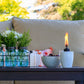 White ceramic oil lamp with flame, set on outdoor table with flowers and bowl of cherries.