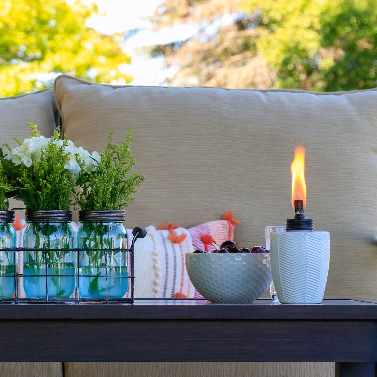 White ceramic oil lamp with flame, set on outdoor table with flowers and bowl of cherries.