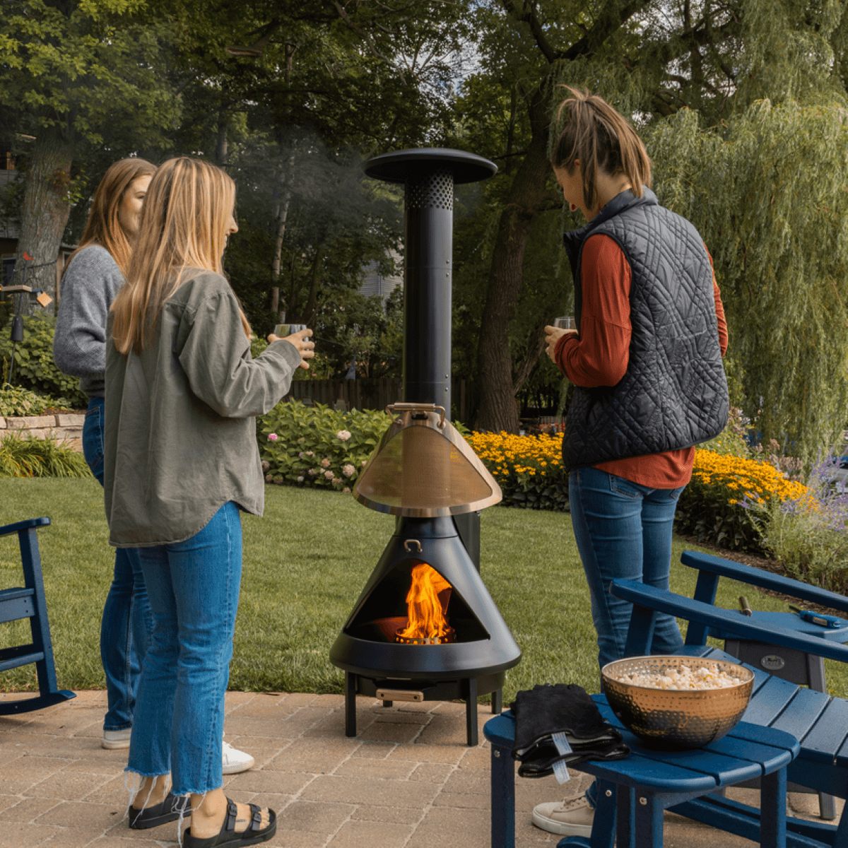 Black outdoor chiminea fireplace with flame, surrounded by people and patio furniture.