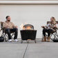 Black fire pit with flames, two people sitting in chairs around it.