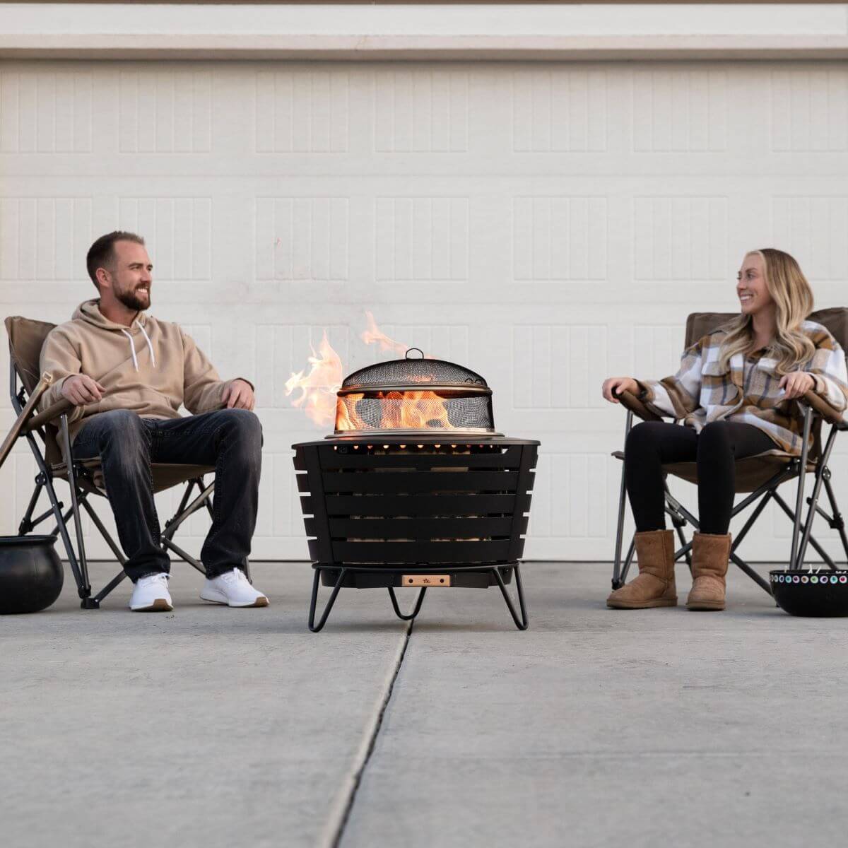 Black fire pit with flames, two people sitting in chairs around it.