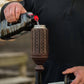 Dark brown Tiki torch with circular cutouts, being filled with fuel from a bottle.