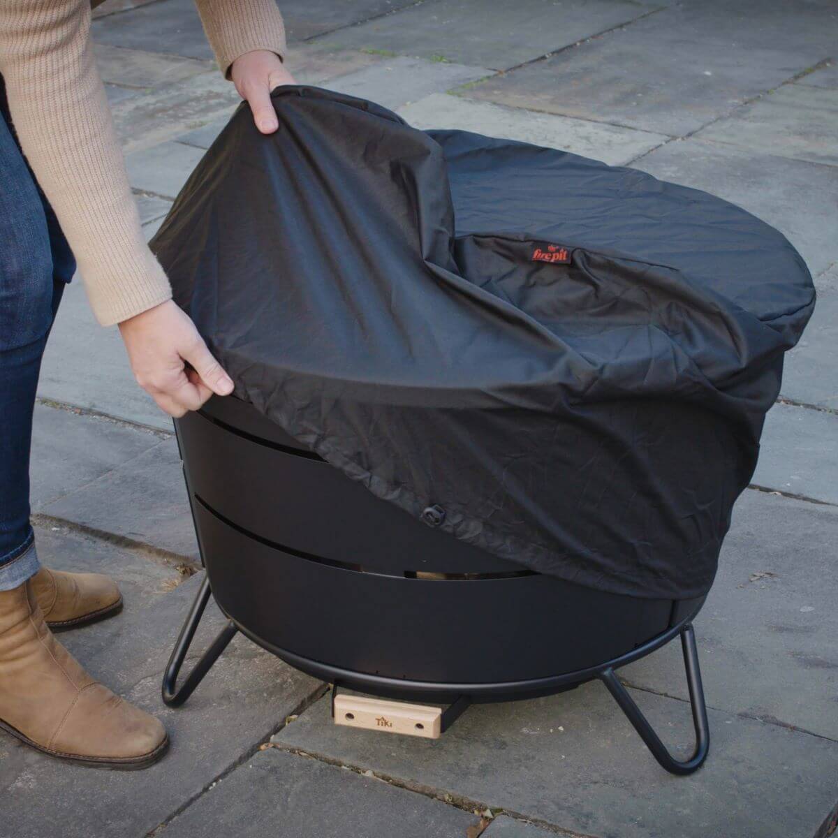 Black fire pit cover being placed on a round fire pit with legs.