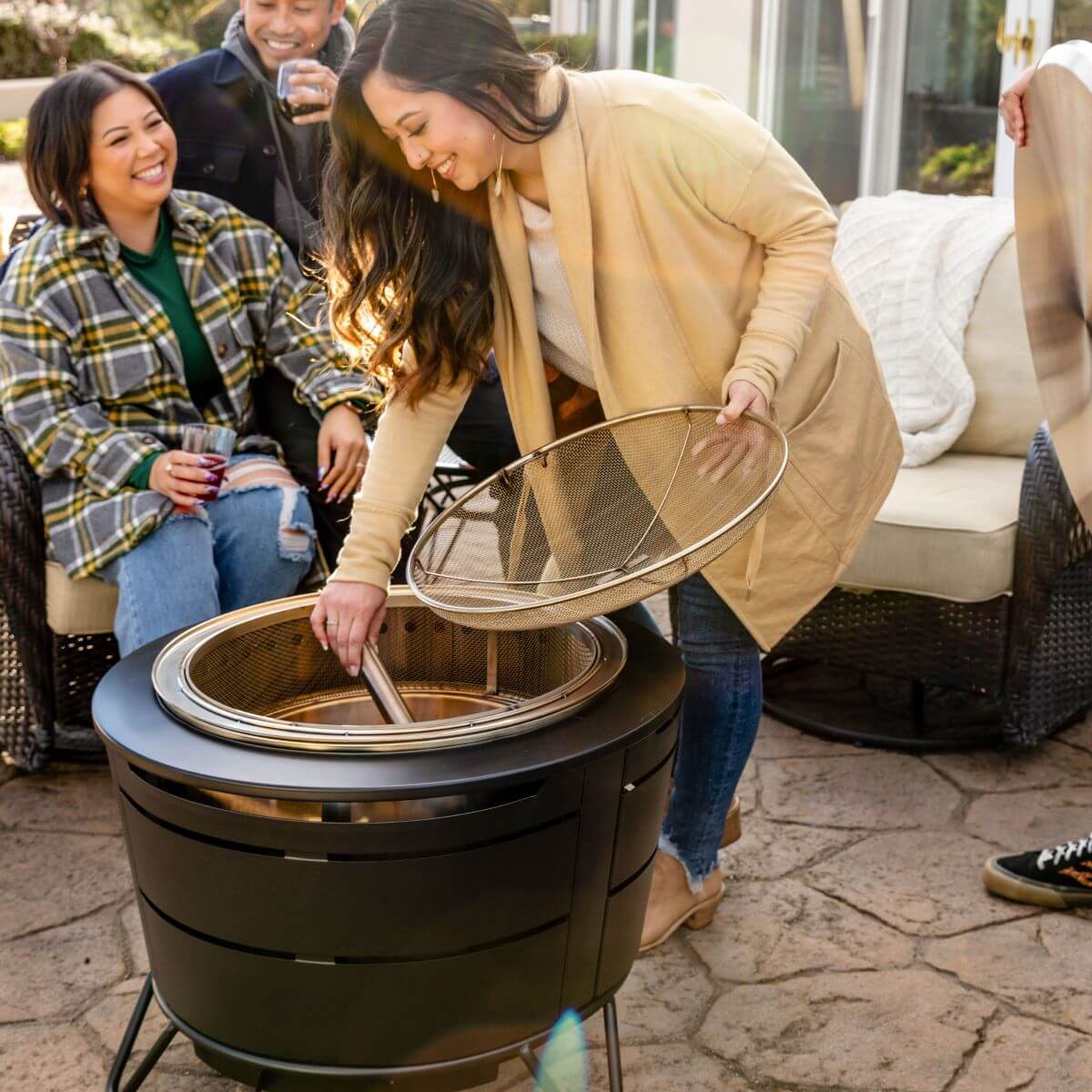 Black outdoor fire pit with mesh lid and tools, surrounded by people enjoying drinks.