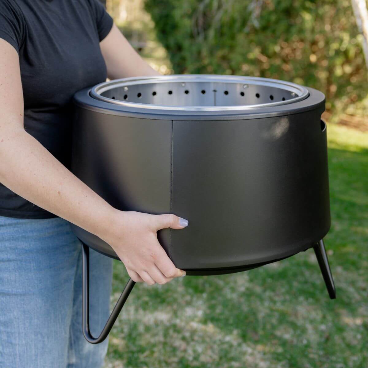 Black portable charcoal grill with lid, held by person outdoors.