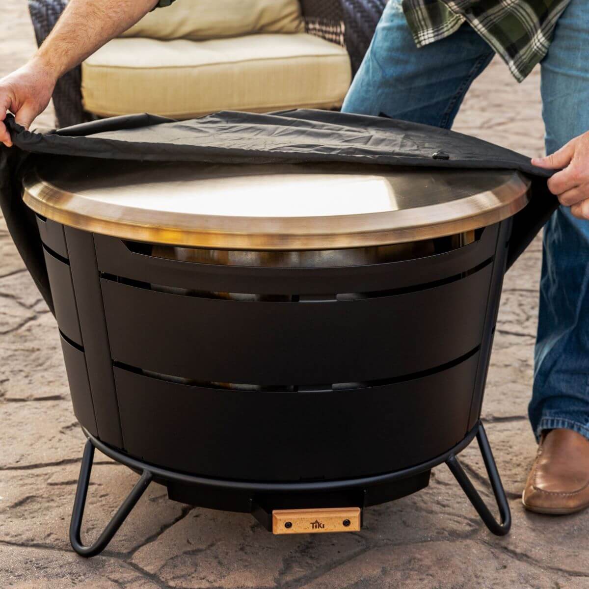 Black outdoor fire pit with gold lid and protective cover being placed on top.