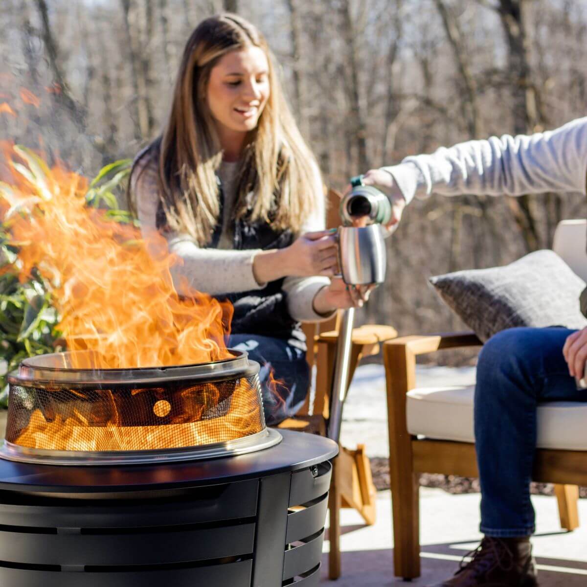 Black fire pit with flames, outdoor seating, and people enjoying coffee.