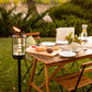 Black outdoor torch with flame, set on grass near wooden table with bread, plates, and glasses.